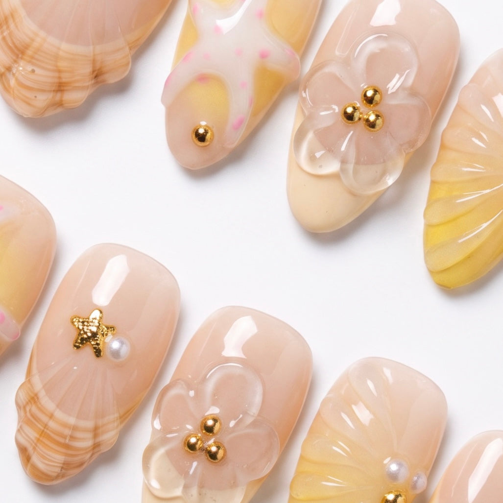 Close-up of handmade seashell press-on nails featuring sculpted shell textures, tiny flowers, pearls, and gold embellishments.
