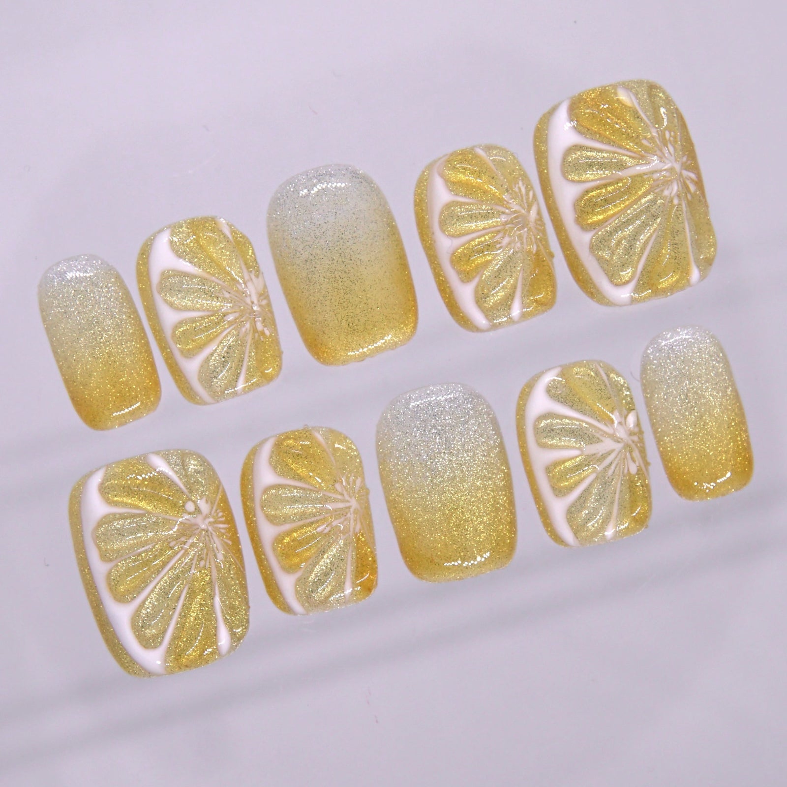 Close-up of yellow shimmer press on nails featuring white outlined citrus-inspired artwork and fine glitter gradient