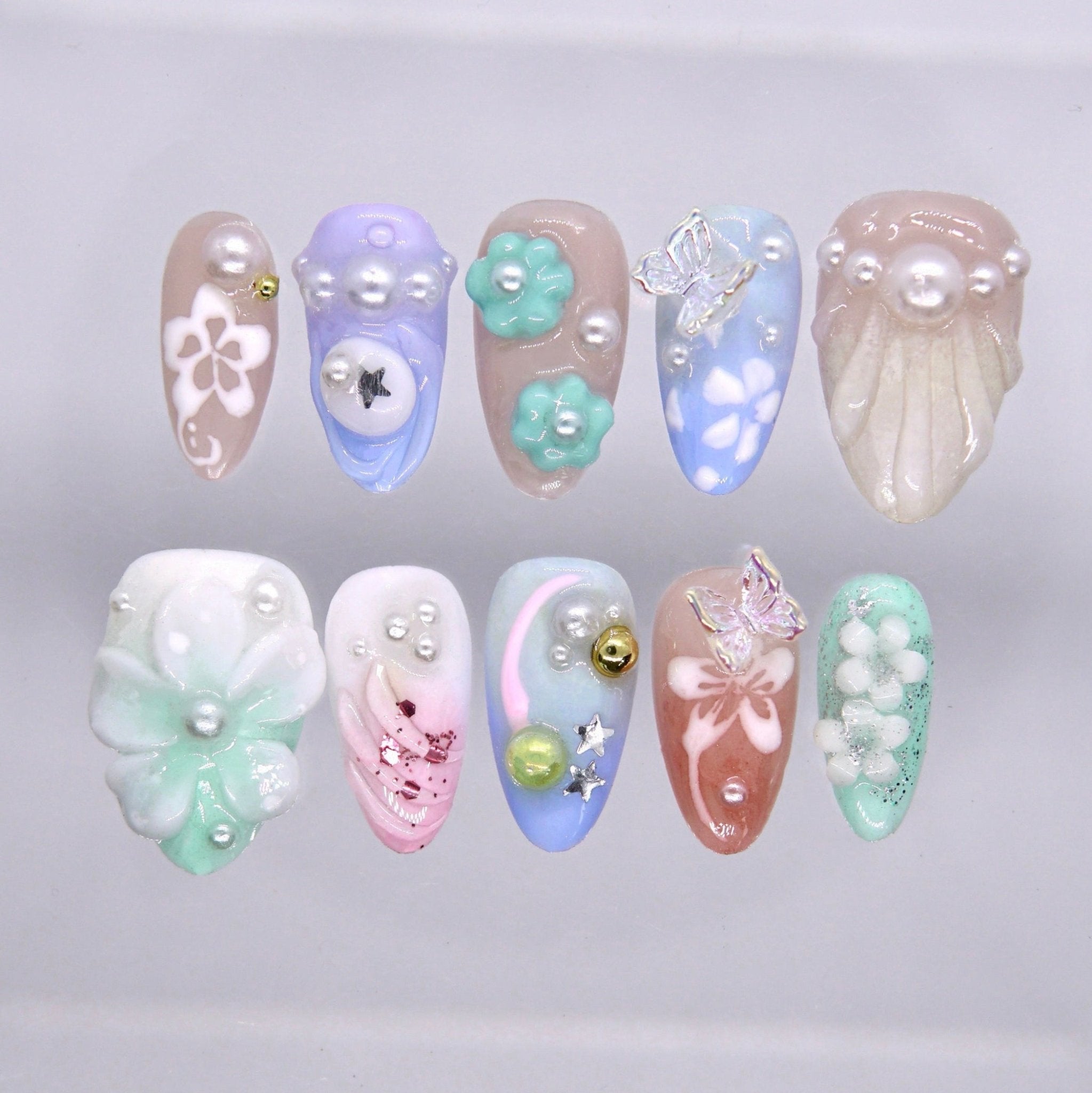 Long almond press on nails with pastel blue, mint, blush, and lavender tones featuring flowers, pearls, butterflies, and sculpted charm details