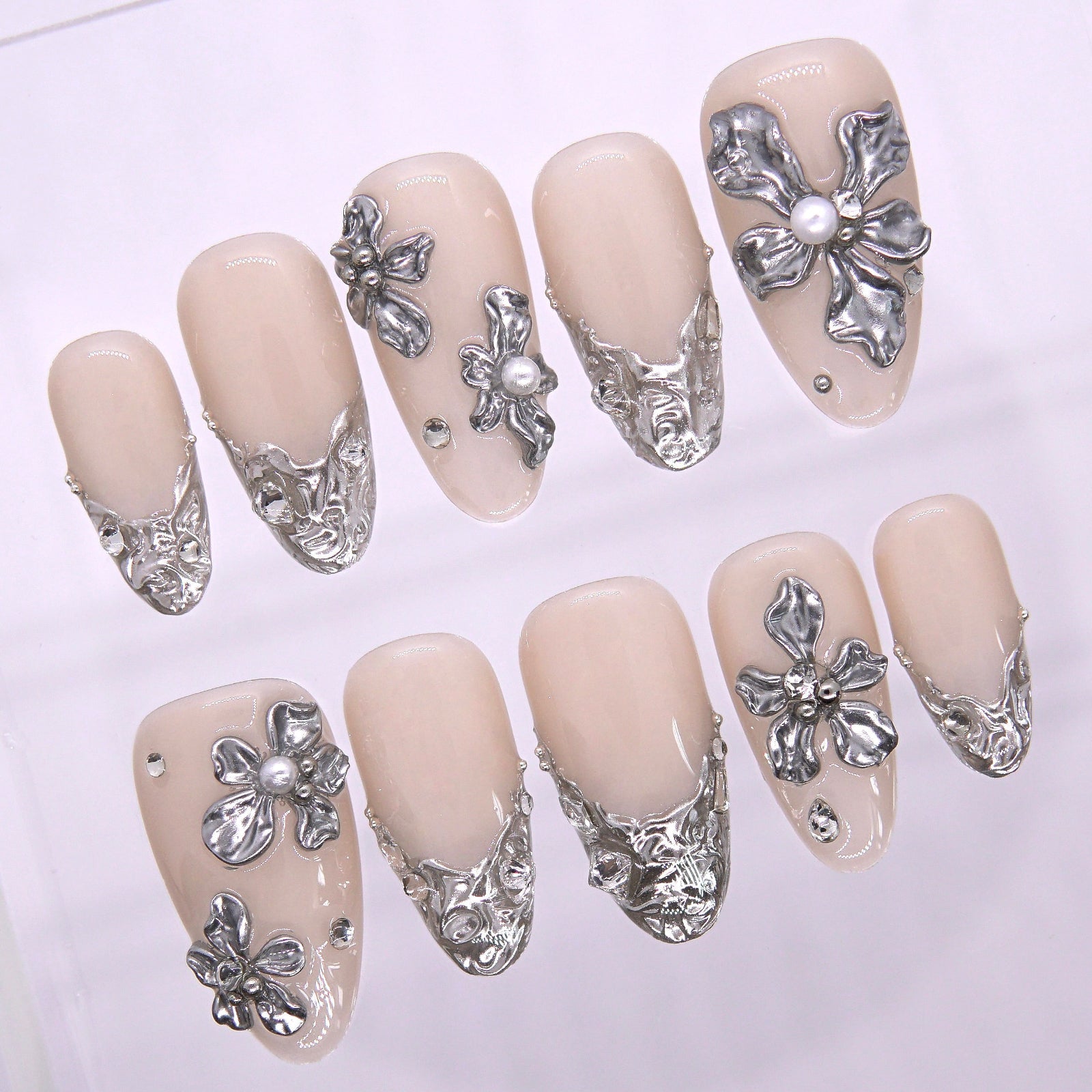 Close-up of large metallic silver floral embellishment on nude nail surface, showcasing detailed carving and pearl-like structure.