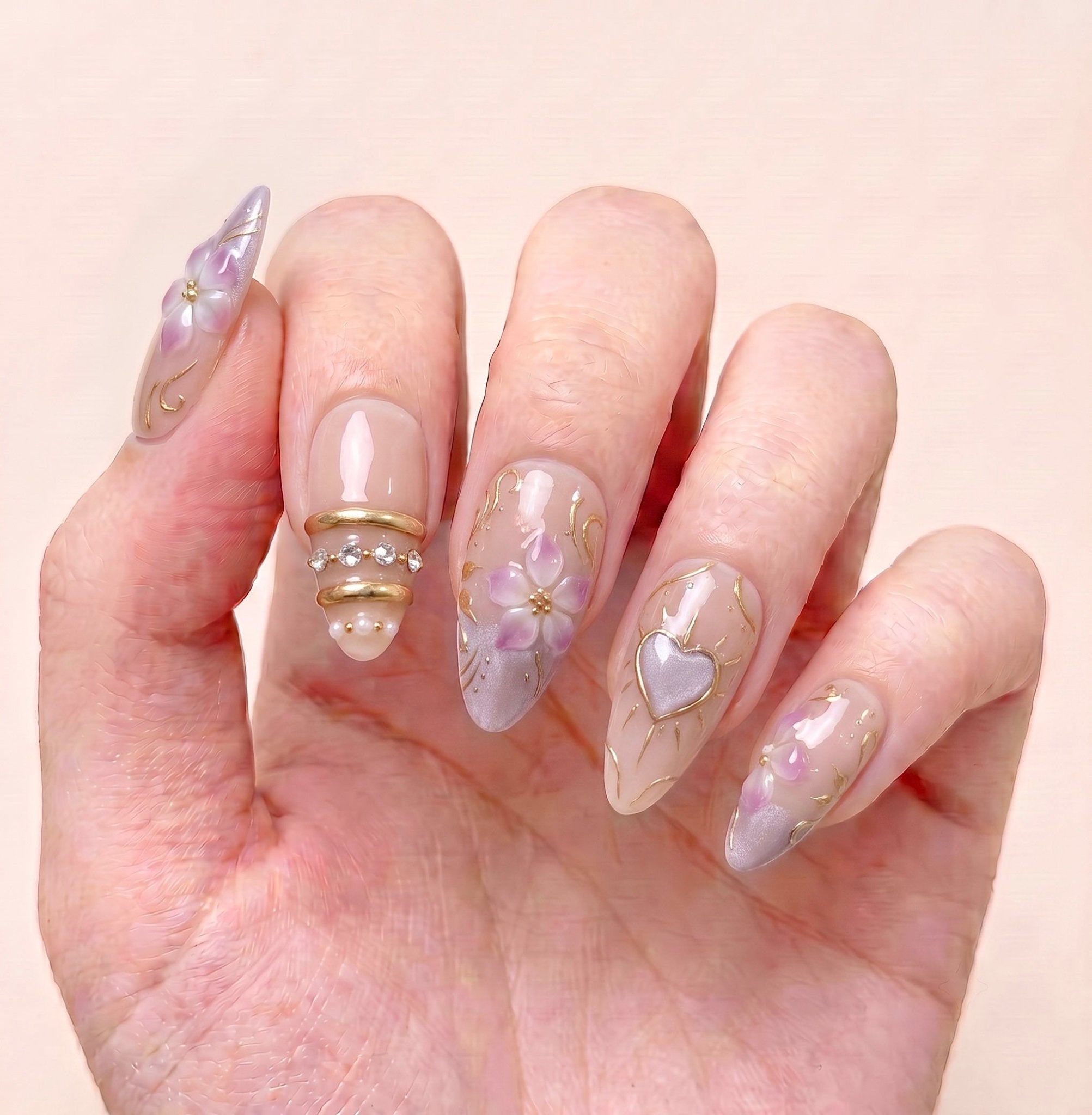 Heartspell Garden handmade press-on nails worn on hand with soft nude tones, lilac flowers, gold vine details, hearts, and crystals