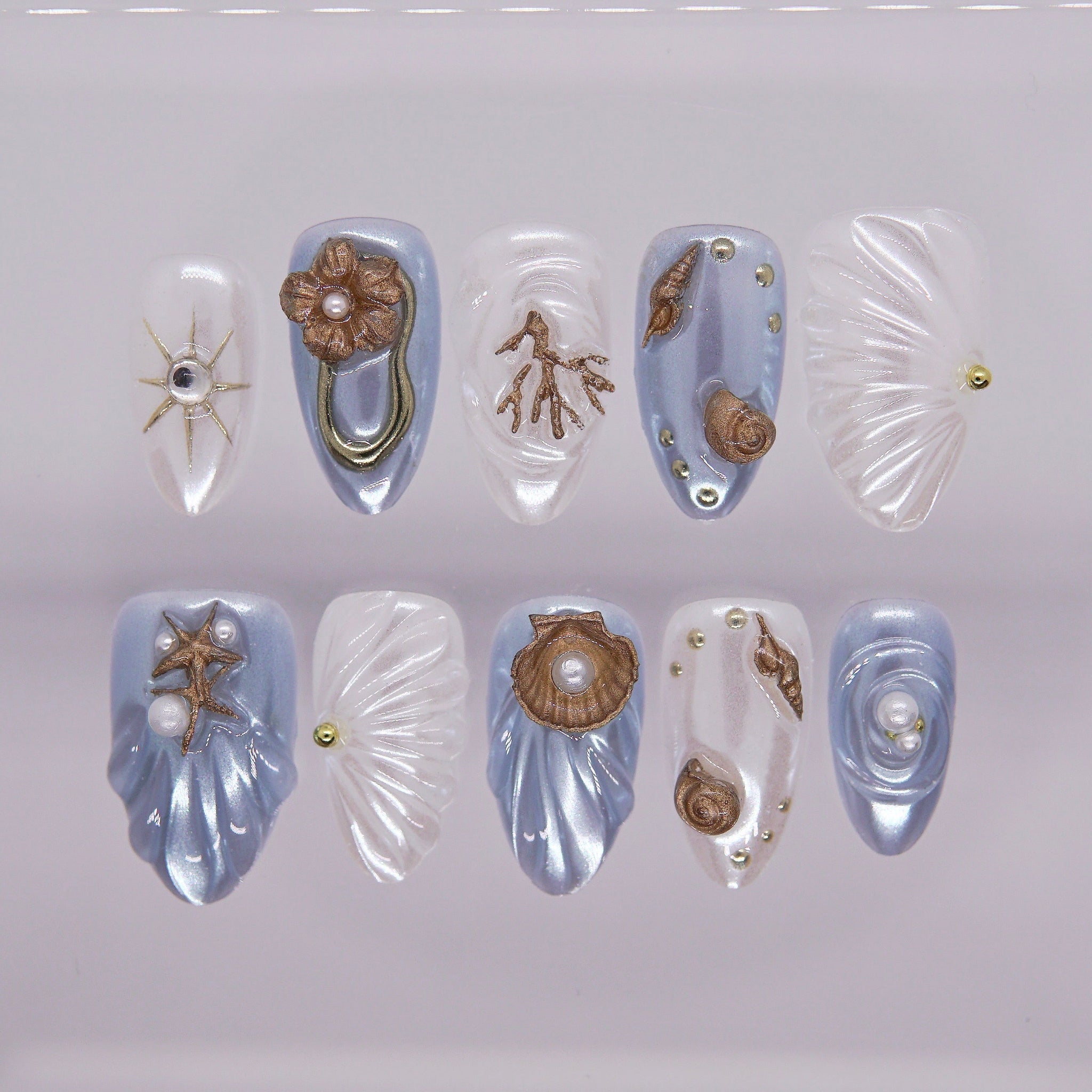 Press on nails with sculpted seashell design in pearl white and blue, decorated with bronze starfish, coral, and pearls. 